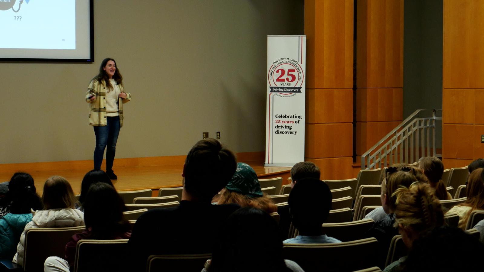 MCDB Student Emily Payne presenting on stage during the 2025 MCDB 3MT competition, as students watch from the audience